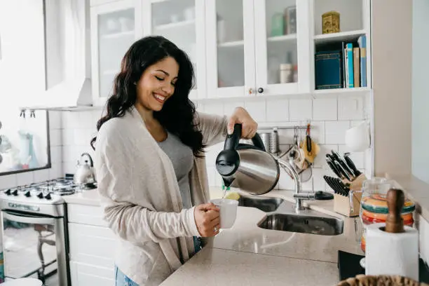 Young adult woman filling a cup of coffee. She's smiling. Hispanic ethnicity.
