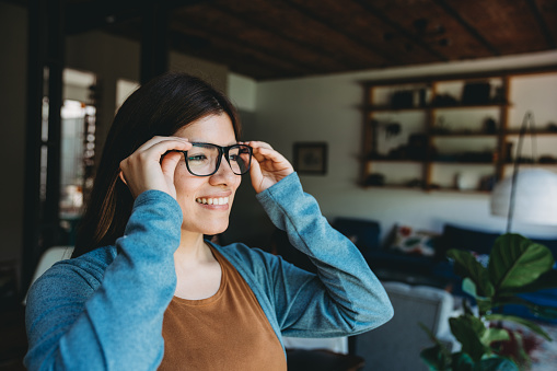 Young adult woman is cleaning her eyeglasses near a window at home. Hispanic ethnicity.