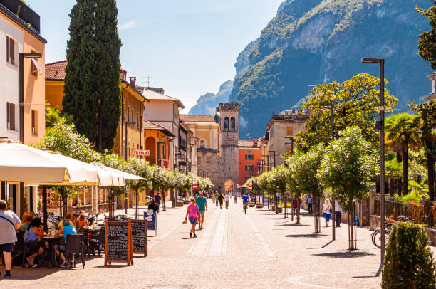 scenic cityscape of riva del garda. cozy city street full of tourists, plants and italian architecture with high dolomite mountains on background - tourist resort lake italy scenics imagens e fotografias de stock