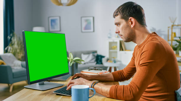 istock Handsome Smiling Man Sitting at His Desk at Home Uses Personal Computer with Mock-up Green Screen. He Works in Cozy Aparment with Modern Living Room Interior. 1196367877