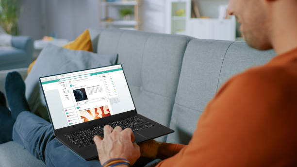 Handsome Young Man Sits at Home Uses Laptop and rowses Through Social Network Feed Site. Over The Shoulder Shot of Anonymous Man in the Living Room.