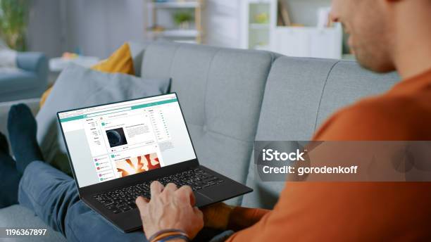 Handsome Young Man Sits At Home Uses Laptop And Rowses Through Social Network Feed Site Over The Shoulder Shot Of Anonymous Man In The Living Room Stock Photo - Download Image Now