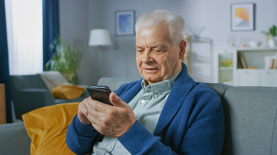 Portrait of Progressive Senior Man Sitting in His Living Room Easily Uses Smartphone, Does Touching Gestures and Feels Very Comfortable with New Technologies.