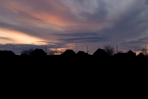 The silhouette of houses and roofs against the sunset. Rural houses and trees on the background of a colorful gorgeous sunset.
