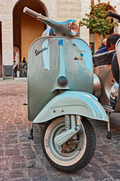 Elderly man riding a vintage black scooter through a narrow cobblestone street in the historic center of Naples, Italy. The surrounding alley features traditional buildings, laundry hanging from balconies, potted plants, and vibrant urban life. A typical southern Italian cityscape depicting everyday life and local culture.