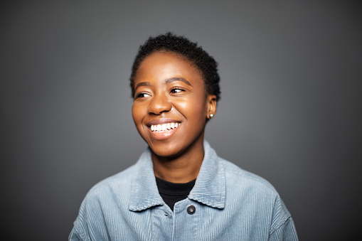 Shot of happy young woman standing against grey background. Happy african american female in denim shirt looking away smiling.
