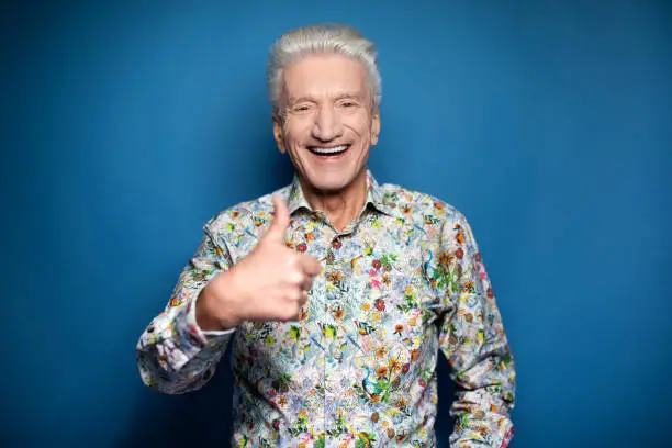 Portrait of a senior man in colorful floral printed shirt giving thumbs up and looking happy. Cheerful mature man gesturing ok sign against blue background.