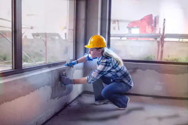Beatiful woman fixing the wall in new home.Female with hard hat working in renovation house. Fixing wall surface and preparation for painting, home renovation concept.