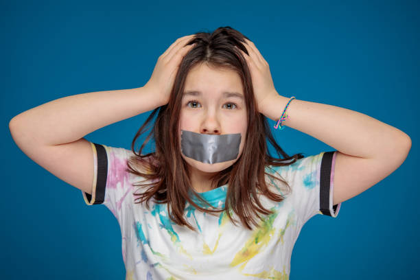 noisy teenager girl silenced with a mouth sealed with silver tape and head in hands stock photo