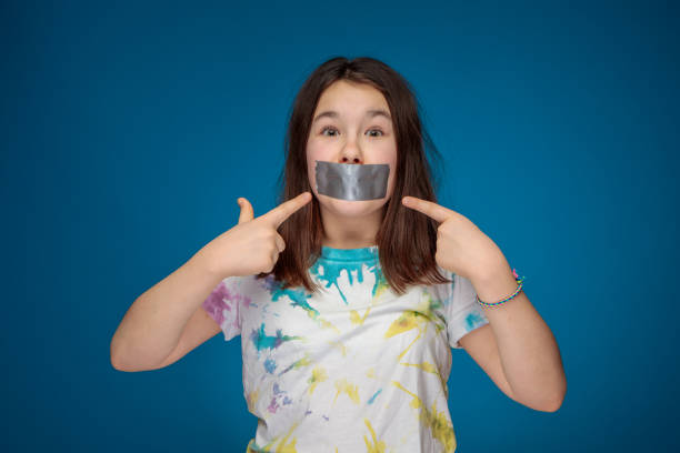 noisy teenager girl silenced with silver tape pointing at her mouth A noisy teenage girl in a self-made colorful t-shirt silenced with silver tape. Studio shot on blue background. speaking with forked tongue stock pictures, royalty-free photos & images