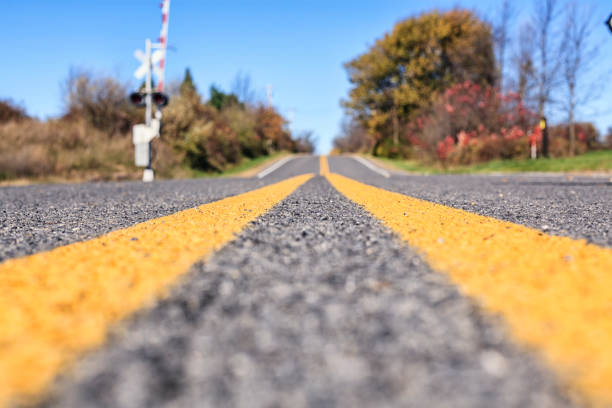 Extreme selective focus surface level view of a rural, gray asphalt country road, with bright yellow unbroken dividing line road markings near the intersection of the highway with a rarely-used, but automated warning sign equipped railroad crossing. Photo taken in western New York State, about 20 miles southeast of the city of Rochester, NY.