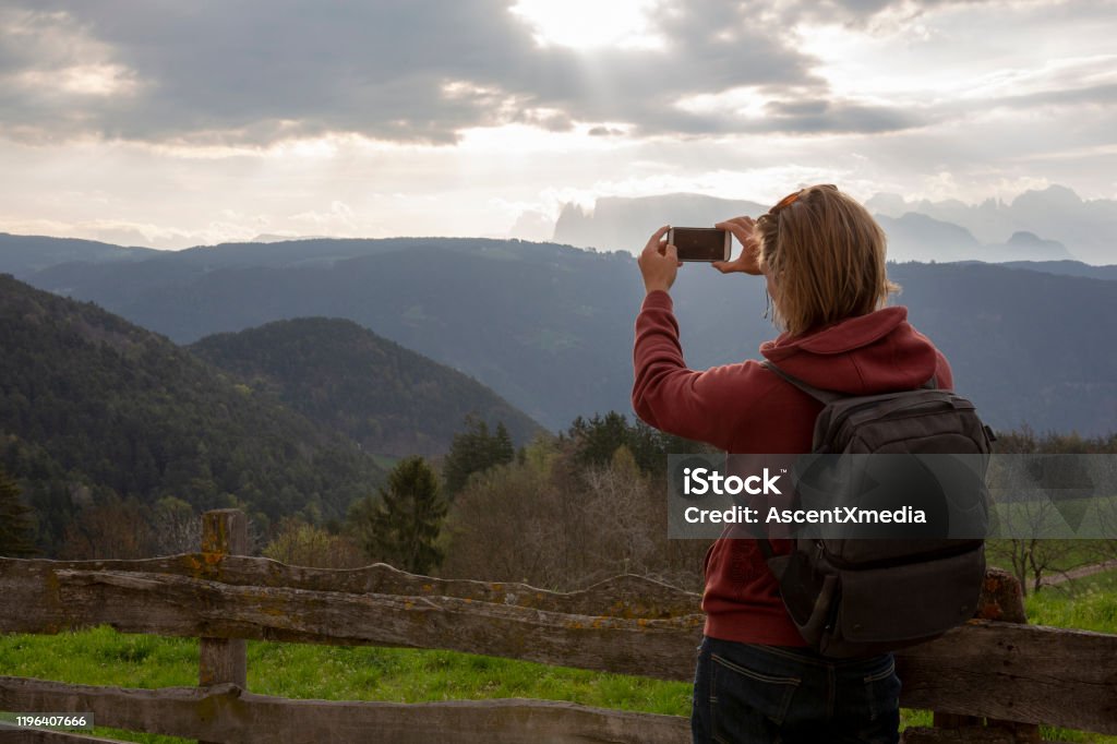 Young man leans on wooden fence in the hills and takes photo He looks off to distant scene Blond Hair Stock Photo