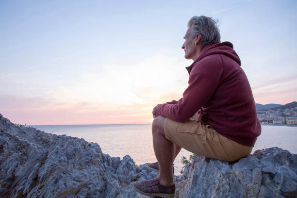 成熟した男は日の出時に海の上の岩の尾根に座っている - 15824 ストックフォトと画像