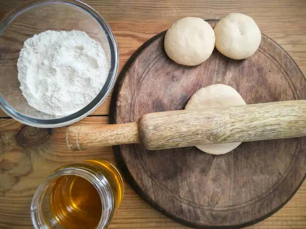 Rolling pin and small bolls of wheat dough with flour, oil on wooden table.