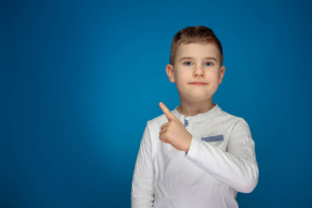 A small boy in white long sleeve pointing with a finger over his shoulder. Studio shot on blue background.