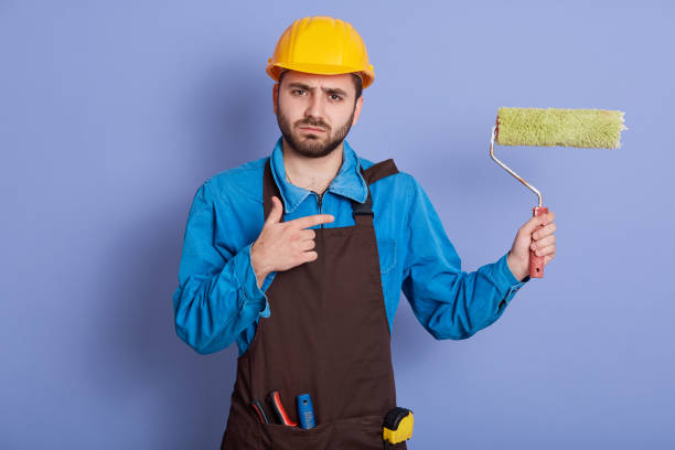 istock Image of bearded male builder going to paint walls, pointing at paint roller with his index finger, wearing special uniform and protective helmet. Flat renovation, painting and building concept. 1196507801
