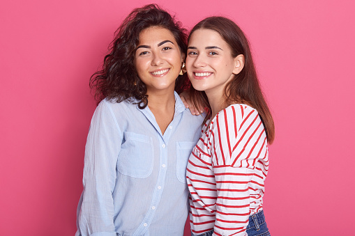 Lovely Caucasian females embracing and smiling to camera, posing isolated over pink background, dresses stylish shirts, attaractive girls expressing love to each other. One sexl ove concept.