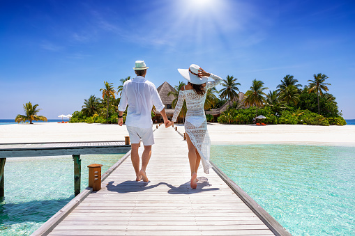 A happy traveler couple in white summer clothing walks down hand in hand a wooden pier towards a tropical paradise island in the Maldives, Indian Ocean