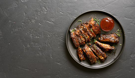 Spicy hot grilled spare ribs on plate over black stone background with copy space. Tasty bbq meat. Top view, flat lay