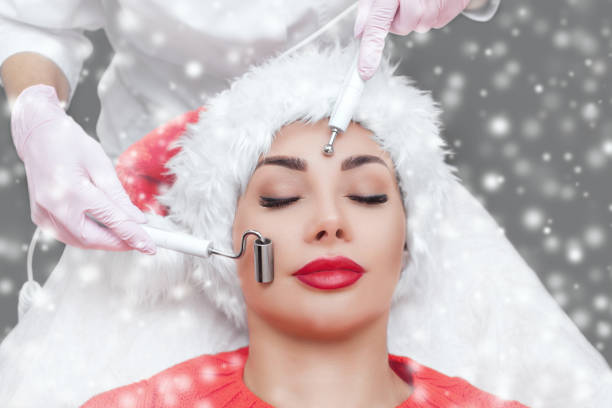 o cosmetologista faz o procedimento da terapia microcorrente de uma mulher no chapéu de papai noel em um fundo da neve. conceito de ano novo e cosmetologia. - galvanic - fotografias e filmes do acervo