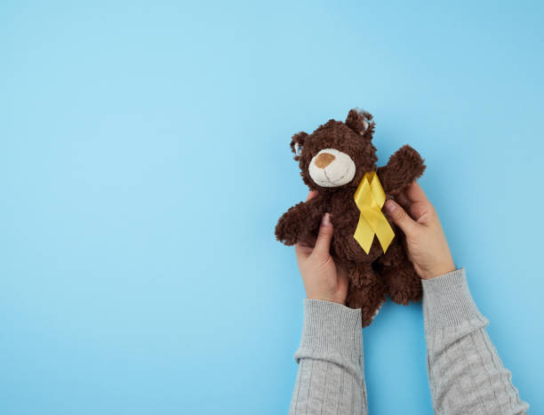 female hands hold a small brown teddy bear which holds in its paw a yellow ribbon female hands hold a small brown teddy bear which holds in its paw a yellow ribbon folded in a loop on a blue background. concept of the fight against childhood cancer. problem of suicides and their prevention behavior teddy bear doll old stock pictures, royalty-free photos & images