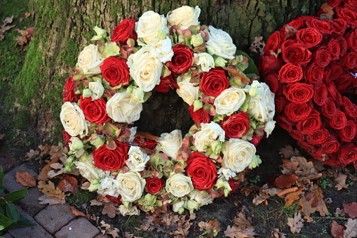 A classic funeral wreath made of red and white roses near a tree, surrounded with fallen autumn leaves.