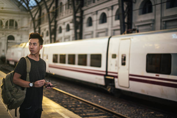 Hipster Chinese man with backpack waiting for the train on the railway station and texting on the mobile phone