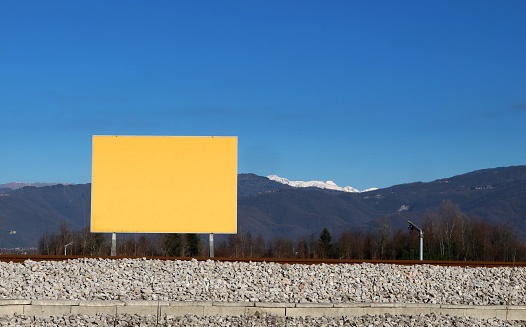 Empty yellow advertising billboard on a stone embankment in countryside. Mountains and blue sky on background. Copy space