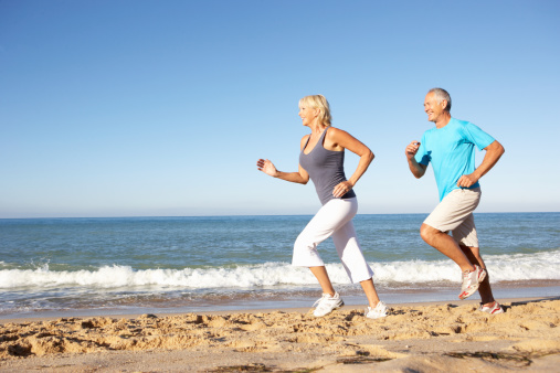 Senior Couple In Fitness Clothing Running Along Sandy Beach Together