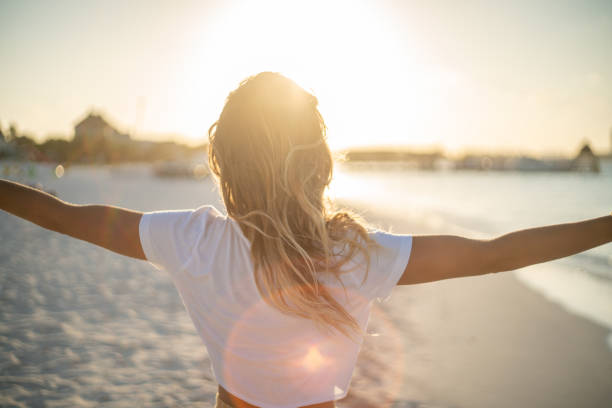 Cheerful young woman embracing nature at sunset; female standing on beach arms outstretched stock photo