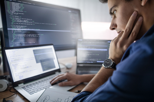 A focused young man with a watch is sitting at a desk, intently examining code on multiple computer screens and a laptop, with programming graphics and lines of text visible on the displays.