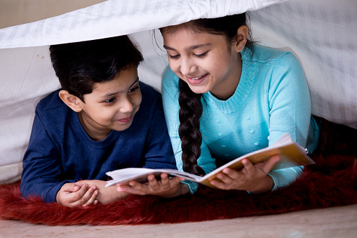 Happy boy and girl reading a book under blanket at home