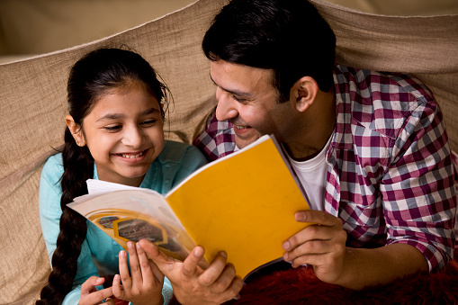 Happy father reading a book to his daughter under blanket at home
