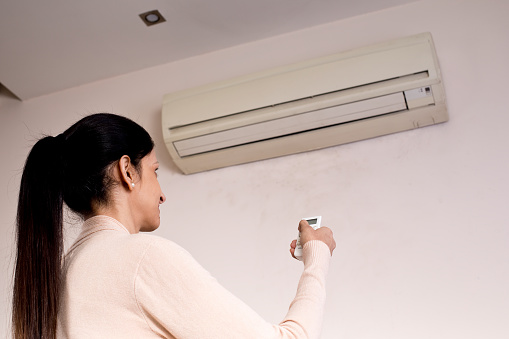 Woman switching on or adjusting the temperature of wall mounted air conditioner with a remote control at home
