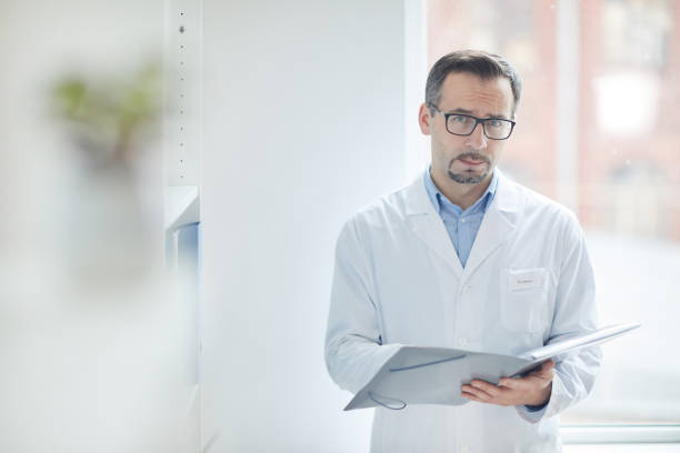 Doctor filling the medical card Portrait of mature male doctor in white coat standing and looking at camera while filling the medical card at office technology office equipment laboratory stock pictures, royalty-free photos & images