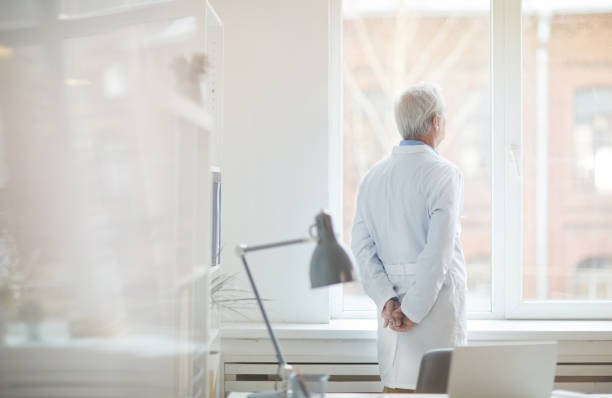 Rear view of senior male doctor in white coat standing near the window at office thinking and resting after work