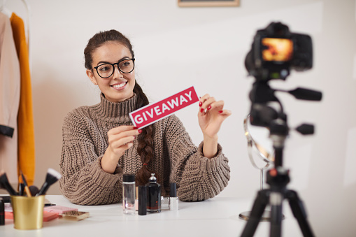 Portrait of contemporary young woman holding GIVEAWAY word while filming video for beauty and lifestyle channel, copy space
