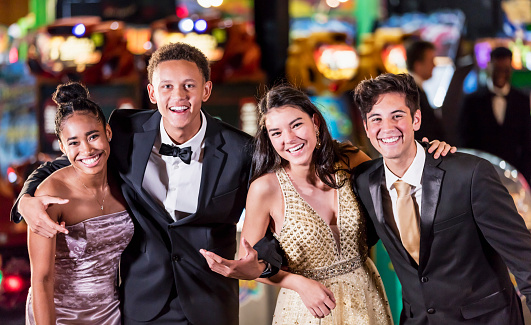 A multi-ethnic group of four teenagers, two interracial couples, having fun at their high school prom. The two girls are wearing prom dresses and their dates are wearing a suit and tuxedo.