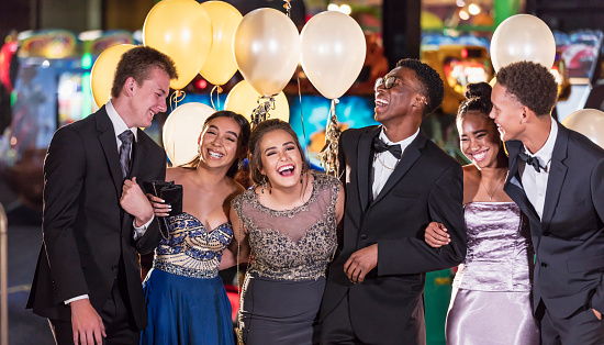 A multi-ethnic group of six teenagers, three multi-ethnic couples, having fun at their high school prom. The two girls are wearing prom dresses and their dates are wearing a suit and tuxedo.