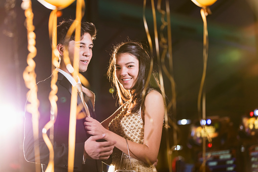 A multi-ethnic teenage couple having fun at the prom. They are both 17 years old. They are indoors at night, with balloons out of focus around them, on the dance floor. The Hispanic teenage boy is wearing a suit and tie, and his date is wearing a prom dress.