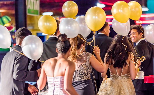 Rear view of a group of multi-ethnic teenagers at prom. They are wearing prom dresses and tuxedos, indoors at night, holding gold and white balloons.