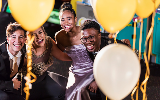 A multi-ethnic group of four teenagers, two interracial couples, having fun at their high school prom. The two girls are wearing prom dresses and their dates are wearing a suit and tuxedo. They are laughing and looking at the camera through balloons in the foreground.