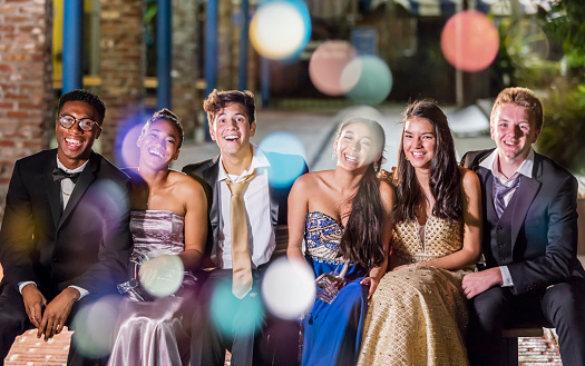 A multi-ethnic group of six teenagers, three multi-ethnic couples, having fun hanging out after their high school prom. The girls are wearing prom dresses and their dates are wearing a suits and tuxedos. Some of the girls are holding their shoes and the guys' ties are untied.