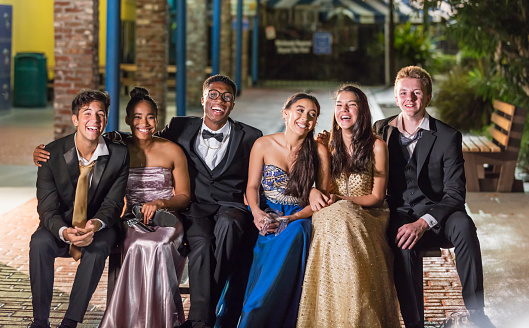 A multi-ethnic group of six teenagers, three multi-ethnic couples, having fun hanging out after their high school prom. The girls are wearing prom dresses and their dates are wearing a suits and tuxedos. Some of the girls are holding their shoes and the guys' ties are untied.