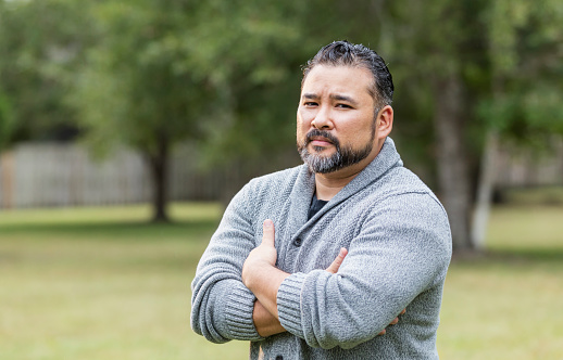 A mid adult Hispanic man in his 30s standing in the park, looking at the camera with a serious expression, arms folded.