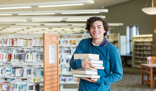 A 16 year old Hispanic teenager, a high school student, standing in a library carrying a large stack of textbooks. He is looking at the camera, smiling.