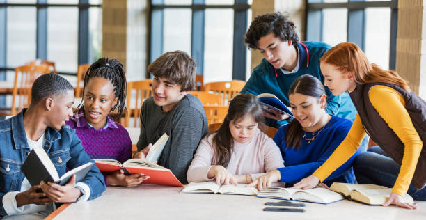 Teenage girl with down syndrome and friends studying A group of seven multi-ethnic teenagers at a table in the library, reading and studying together. The one sitting in the middle wearing the pink hooded shirt has down syndrome. She is 17 years old mixed race Hispanic and Caucasian. She and her friends, teenage boys and girls, 15 to 17 years old, are smiling, looking down at their books, conversing. teenagers only teenager multi ethnic group student stock pictures, royalty-free photos & images