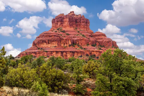 Bell Rock in Sedona on a beautiful sunny day with fluffly white clouds. The landmark is sourrounded by high desert juniper bushes and pines.
