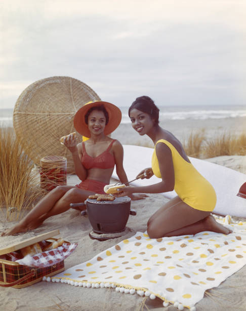 mulheres jovens segurando cachorro-quente na praia, a sorrir - image created 1960s fotos imagens e fotografias de stock