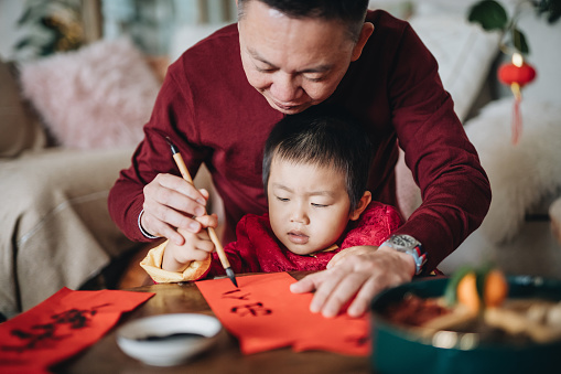 Grandfather practising Chinese calligraphy for Chinese New Year Fai Chun (Auspicious Messages) and teaching his grandson by writing it on a piece of red paper
